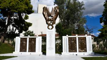 A white monument located in a green park area with a bronze reliefs on the top of it and with engraved names on it.